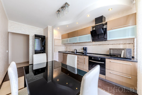 Современная кухня со светлыми фасадами, чёрной столешницей, техникой и обеденной зоной с чёрным стеклянным столом и белыми стульями.