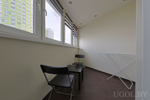 Узкая лоджия с панорамными окнами, двумя складными стульями, небольшим столиком, вентилятором и сушилкой на фоне городских высоток.