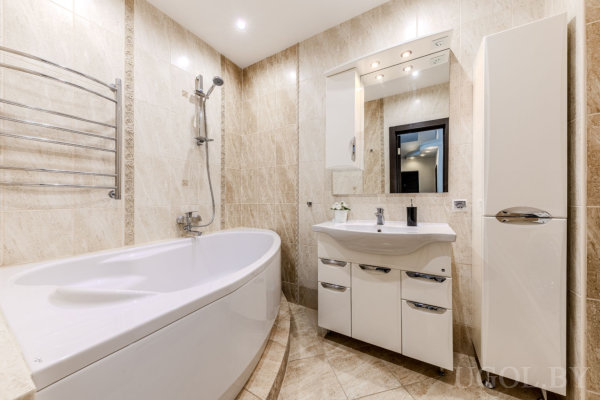 Современная ванная с бежевой мраморной плиткой, большой ванной, тумбой с раковиной, зеркалом с подсветкой и высоким шкафом.