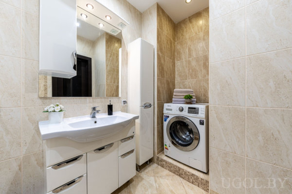 Бежевая ванная с тумбой и зеркалом, высоким шкафом, стиральной машиной с полотенцами и аккуратным современным оформлением.