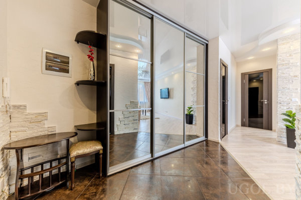 Коридор с большим зеркальным шкафом, декоративной зоной с цветами и табуретом, переходом от тёмной плитки к светлому паркету и акцентной стеной.