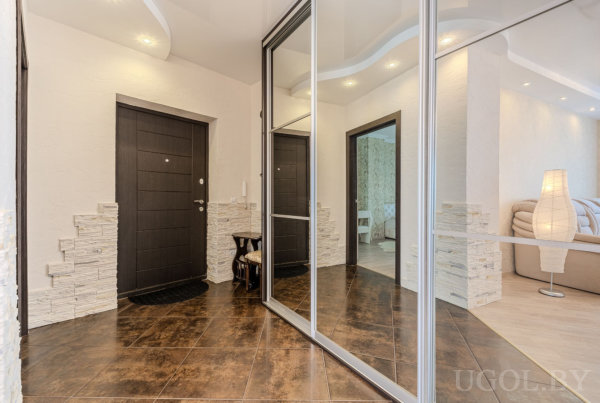 Современный коридор с тёмной входной дверью, зеркальным шкафом, каменной отделкой стен и видом в гостиную с бежевым диваном.