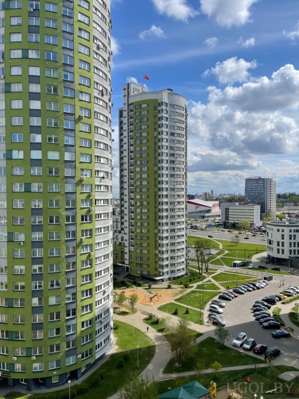 Городской пейзаж с круглыми зелёными башнями, детской площадкой, парковкой и крупным зданием с красно‑белым фасадом на горизонте.