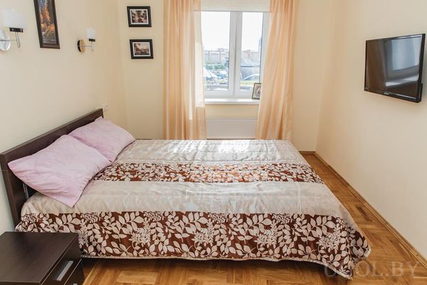 Двуспальная кровать с цветочным покрывалом, прикроватная тумба, картины над изголовьем и окно с тюлем.