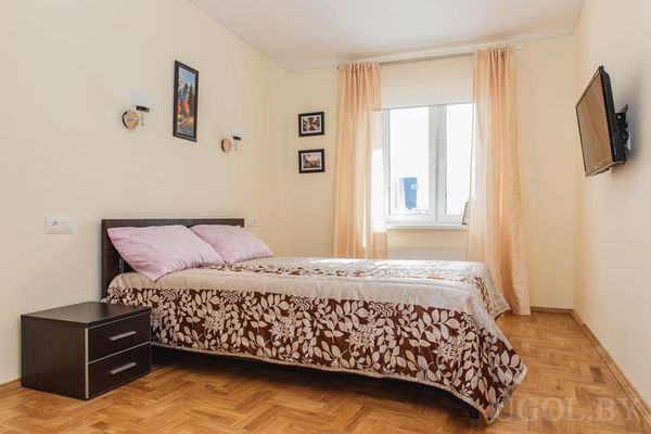 Кровать с коричневым покрывалом, тумба, картины над изголовьем, окно с персиковыми шторами и ТВ на стене.