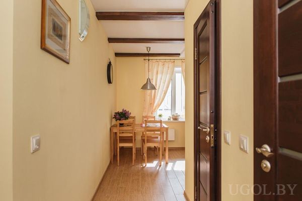 Узкий проход с жёлтыми стенами, деревянным полом и балками, ведущий к столу с цветами у окна.