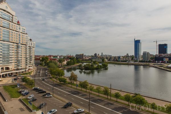 Широкая дорога, высотки, река, стройка с краном, деревья и частично облачное небо.