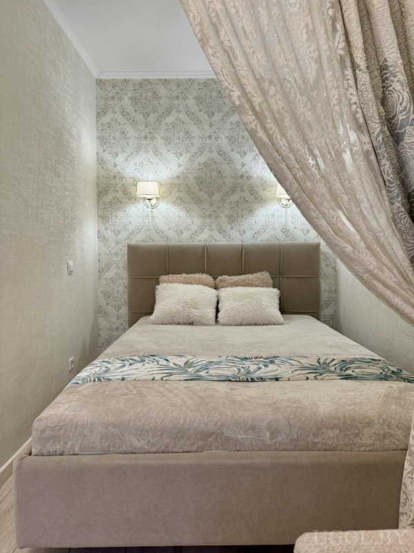 Кровать с вертикальной стёжкой, настенные светильники, кремовые подушки, декоративная дорожка и шторы с цветочным рисунком.