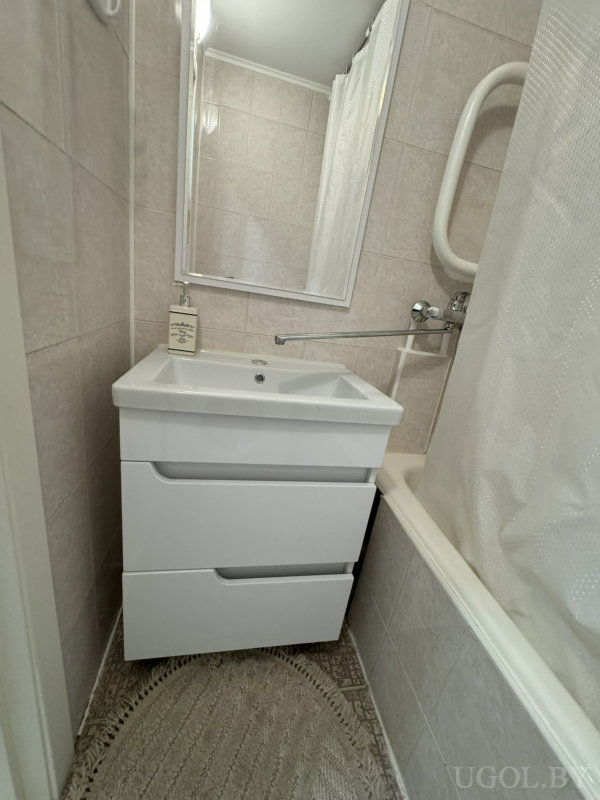 Раковина с зеркалом, белая тумба, мыльный дозатор, ванна с шторкой, настенный душ и полукруглый коврик.