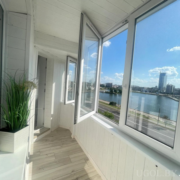 Закрытая лоджия с белой отделкой, деревянный пол, открытое окно, декоративное растение и панорамный вид на город и воду.