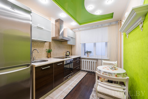 Интерьер с холодильником, плитой, столом с цветочной скатертью, зелёной стеной и растениями.