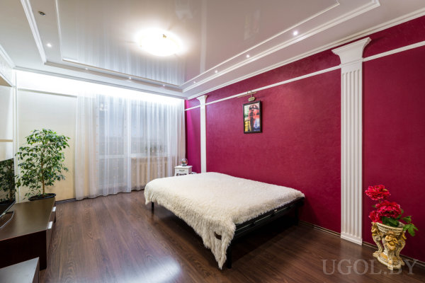 Комната с кроватью, телевизором, вазой с цветами, колоннами на стене и тюлем у окна.