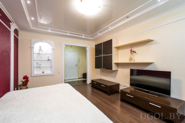 Комната с кроватью, телевизором, полками, нишей с подсветкой и глянцевым потолком.