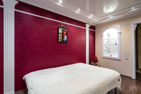 Интерьер с кроватью, декоративной нишей, цветами, картиной и глянцевым потолком с подсветкой.