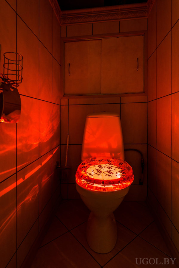 Узкое помещение с унитазом, подсветкой, шкафчиком, ёршиком и плиткой с отражениями.