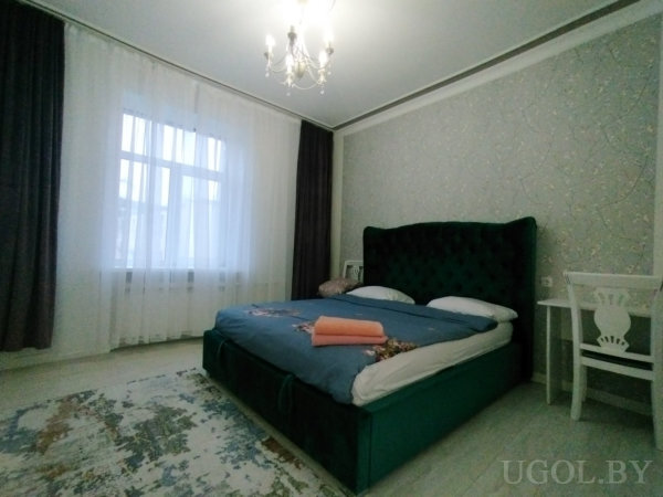 Комната с кроватью, белыми подушками, оранжевым полотенцем, креслом и люстрой.