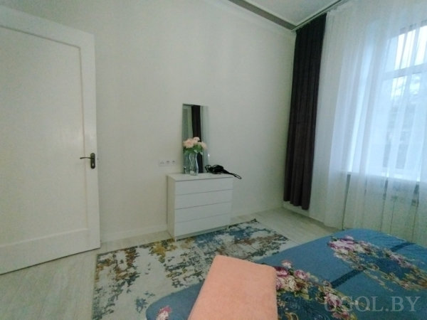 Интерьер с кроватью, комодом, зеркалом, феном, цветами и ковром с орнаментом.