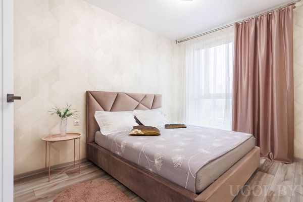 Комната с кроватью в сером текстиле, мягким изголовьем, круглым ковром и вазой с зеленью на прикроватном столике.