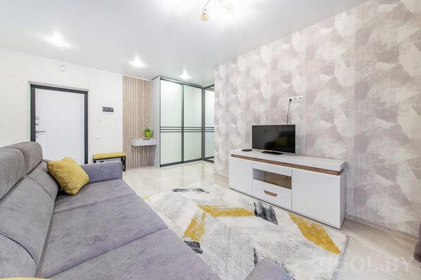 Интерьер с угловым диваном, телевизором на белой тумбе, ковром с геометричным узором и стеклянными перегородками.