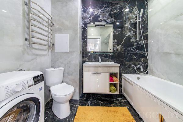 Комната с стиральной машиной, умывальником, ванной и оранжевым ковриком на фоне чёрной мраморной плитки.