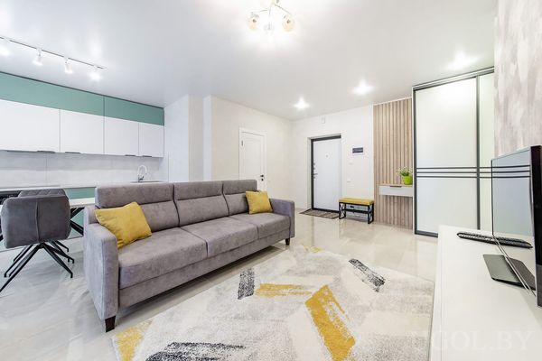 Открытая планировка с серым диваном, ковром с узором, кухней с зелёными фасадами и входной зоной с жёлтой подушкой.