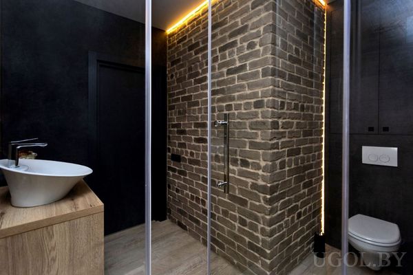 Интерьер с душевой, умывальником, унитазом, тёплой подсветкой и сочетанием дерева, кирпича и тёмных поверхностей.