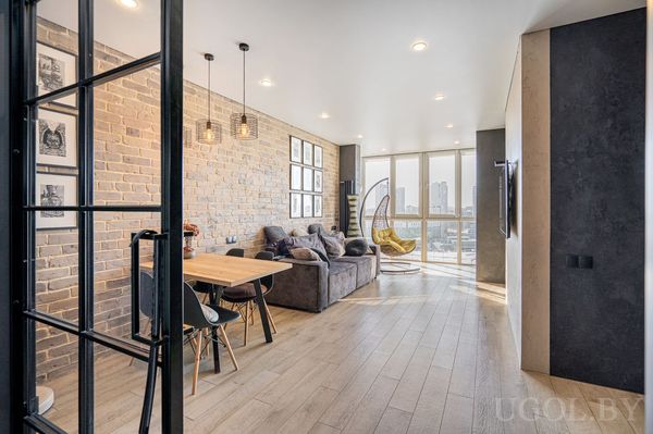 Комната с диваном, креслом, обеденным столом, кирпичной стеной, телевизором и окнами с видом на город.