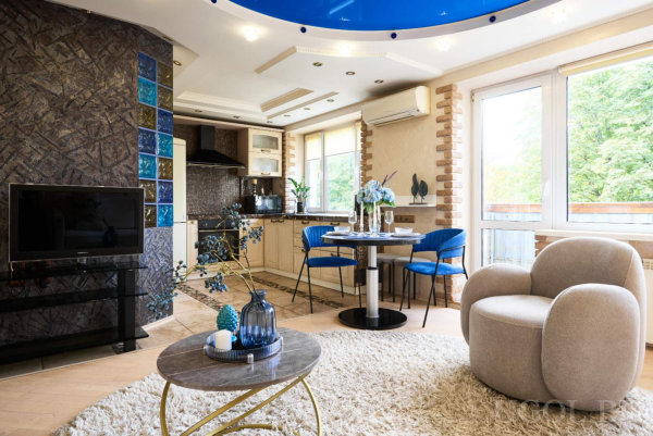 Бежевое кресло на белом ковре, круглый стол с синими стульями, кухня со светлыми шкафами, тёмная декоративная стена с синими стеклоблоками и глянцевый потолок.