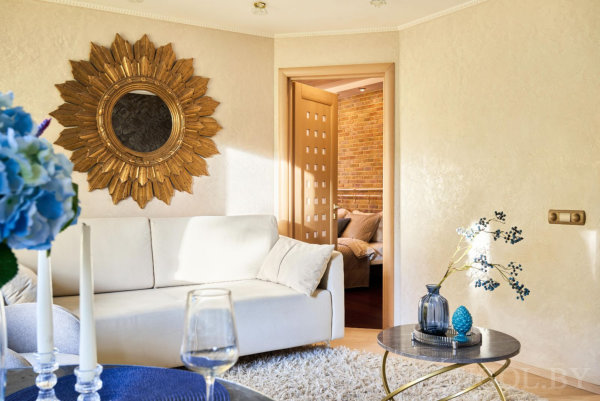 Белый диван у кремовой стены, золотое зеркало‑солнышко, тёмный круглый стол с синим декором, вид в соседнюю комнату с кирпичными акцентами.