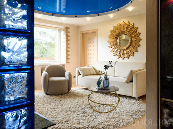 Пушистый кремовый ковёр, серое кресло и белый диван, круглый стол с золотым основанием, синие стеклоблоки и глянцевый потолок с подсветкой.