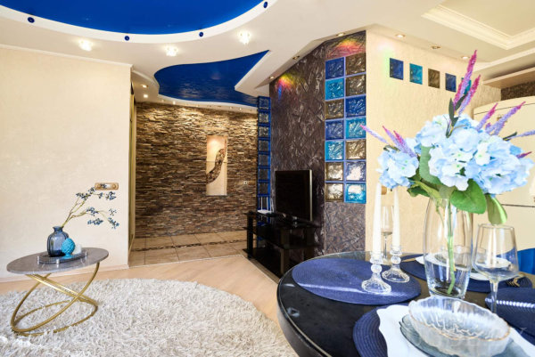 Синий потолок с подсветкой, декоративная стена с цветными стеклоблоками, сервированный стол с цветами, телевизор на тёмной тумбе и белый ковёр.