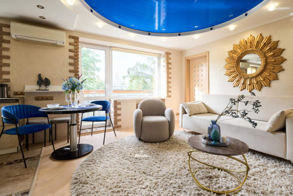 Круглый пушистый ковёр, бежевое кресло и диван, золотой столик, синий потолок, обеденная зона с синими стульями и большим окном.