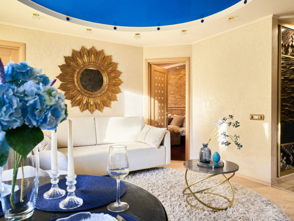 Белый диван у стены с золотым зеркалом, круглый чёрный стол с голубыми акцентами, белый ковёр и вид в соседние комнаты.