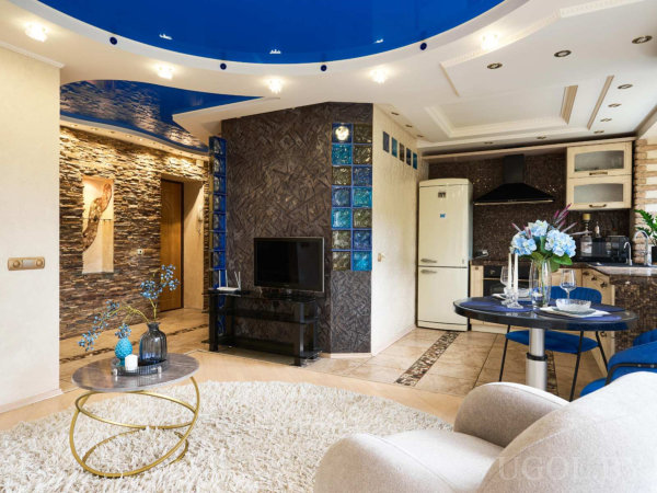 Белый ковёр, стеклянный столик, тёмная стена с синими стеклоблоками, кухня с тёмными шкафами и сервированный стол с цветами.