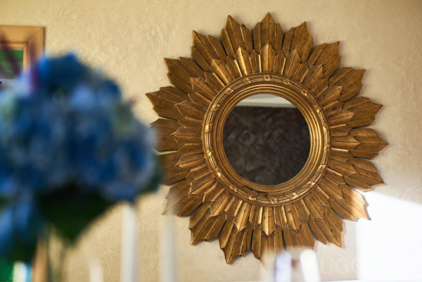 Круглое зеркало с золотыми лучами на бежевой текстурной стене, на переднем плане размытые голубые цветы и свечи.