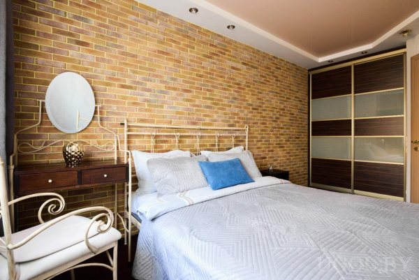 Белая металлическая кровать с голубой подушкой, стена под кирпич, тумбочка с лампой и круглым зеркалом, кресло и шкаф‑купе с тёмными вставками.