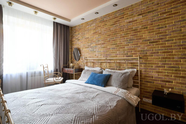 Металлическая кровать с голубой подушкой, кирпичная стена, окно с тюлем и шторами, туалетный столик с зеркалом и тёмная тумбочка.