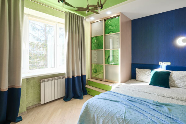 Кровать с серым бельём и бирюзовыми подушками, тёмно‑синяя стена, зелёный потолок с вентилятором, шкаф с зелёными панелями и окно с бежево‑синими шторами.