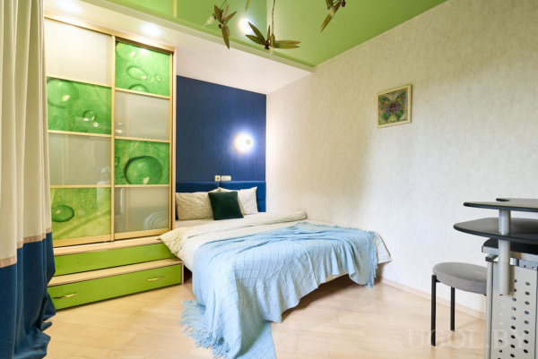 Кровать с бело‑зелёными подушками, тёмно‑синяя стена, шкаф с зелёным рисунком капель, зелёный потолок и столик с серым креслом.