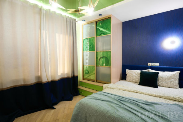 Кровать с бежевым бельём и зелёной подушкой, тёмно‑синяя стена, шкаф с зелёными панелями, зелёный потолок и длинные шторы.