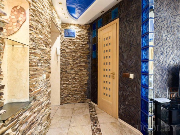 Стена из декоративного камня, дверь со стеклянными вставками, синий глянцевый потолок, вертикальная полоса синих стеклоблоков и каменная дорожка на полу.