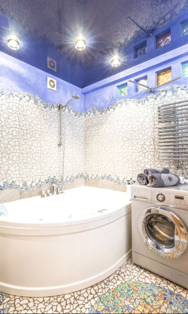 Мозаичные стены и пол, белая угловая ванна с гидромассажем, стиральная машина с полотенцами, полотенцесушитель и синий верх стены.