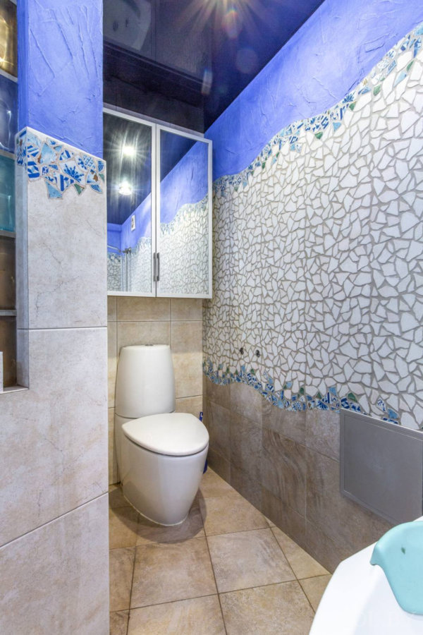 Унитаз у стены с мозаичным узором в виде волн, бежевые плитки, синий верх стены и зеркальный шкаф над унитазом.