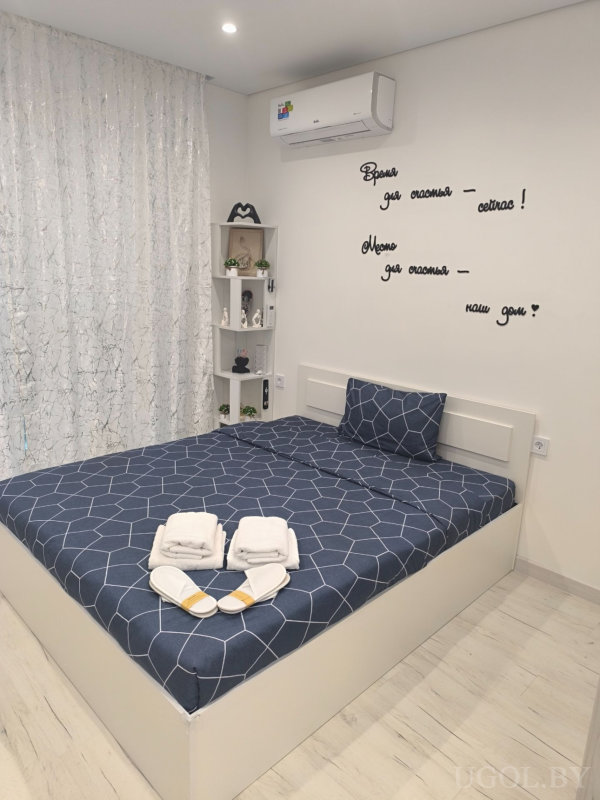 Кровать с синим покрывалом, белыми полотенцами и тапочками, на стене надпись «Время для счастья — сейчас!».