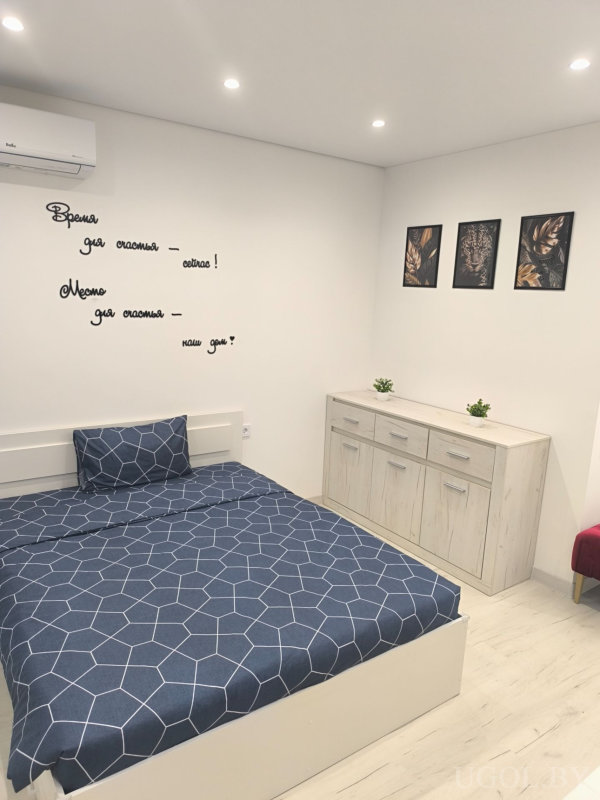 Кровать с синим текстилем, белый комод с зелёными растениями и цитата «Место для счастья — наш дом» на стене.