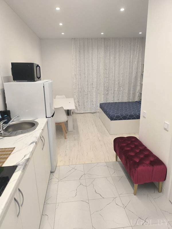 Компактная квартира с кухонной зоной, обеденным столом, кроватью и бордовым пуфом у стены.