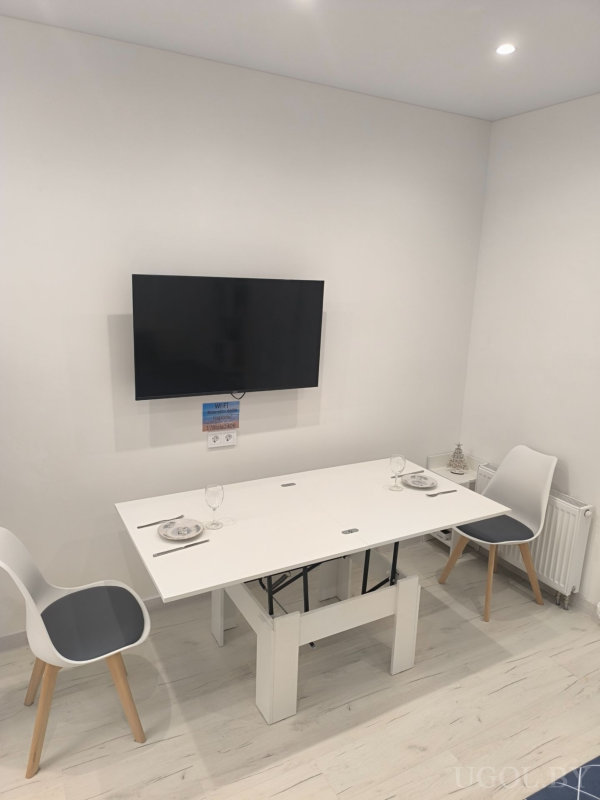 Белый стол на двоих с сервировкой, телевизор на стене и декоративный элемент на батарее.