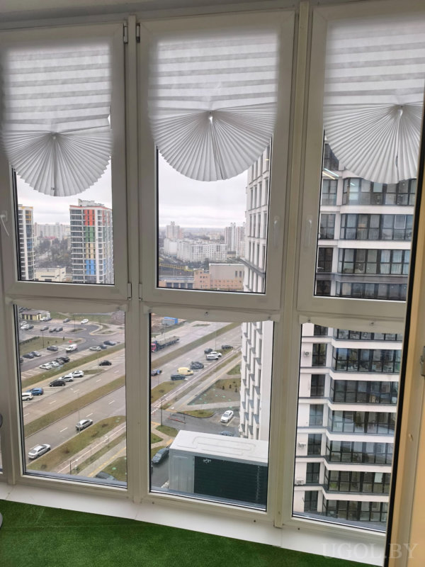 Панорама города с высоты, окна с жалюзи, внизу дорога, высотки и зелёные участки.