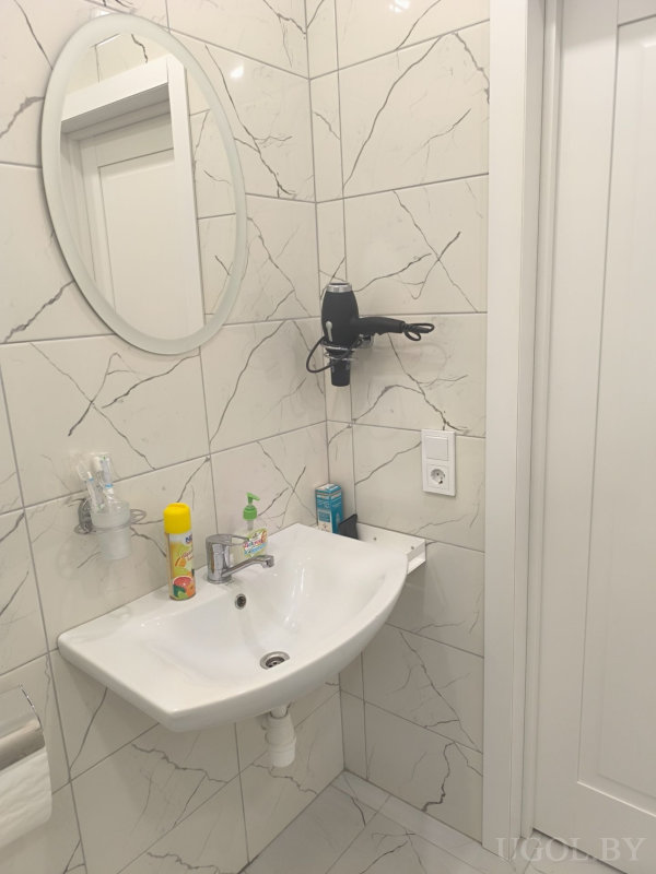 Белая раковина с овальным зеркалом, феном на стене и набором средств гигиены.