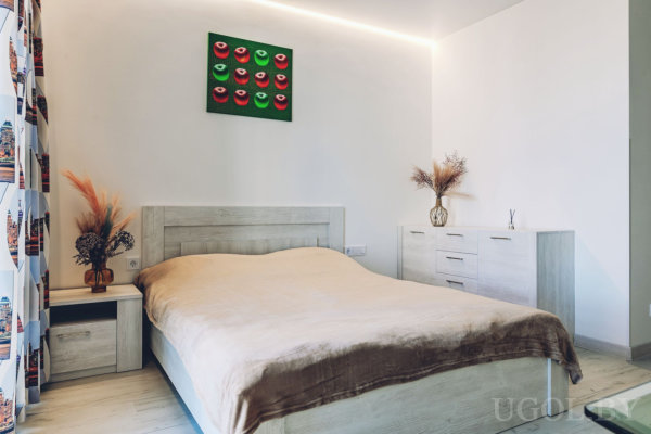 Кровать с бежевым покрывалом и светлыми тумбочками, комод с вазой и декором, белые стены с подсветкой по потолку, на стене картина с яркими лицами в красно-розовой гамме, шторы с городским принтом.